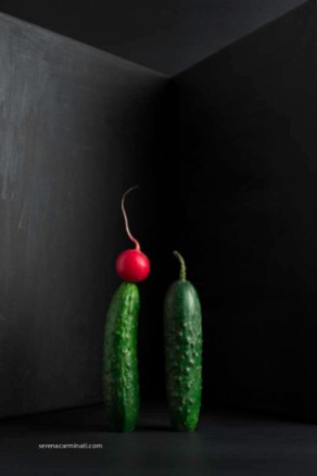 two cucumbers with radish on a dark background, with black painted corner.